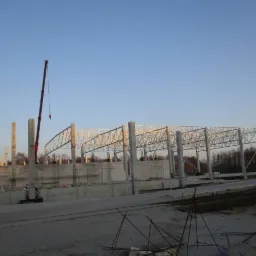 Wczesny etap budowy hali: widoczne betonowe fundamenty, stalowe słupy i konstrukcja dachu, z żurawiem w tle na tle błękitnego nieba o świcie.