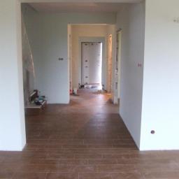 Gerwazbud - Wnętrze w trakcie remontu z brązową podłogą z płytek imitujących drewno, widok na korytarz i surowe ściany z widocznymi instalacjami elektrycznymi.