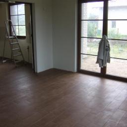 Gerwazbud - Wnętrze pokoju po ułożeniu brązowych płytek podłogowych imitujących drewno, widoczne drzwi balkonowe z powieszonym płaszczem i drabina w przejściu do innego pomieszczenia.