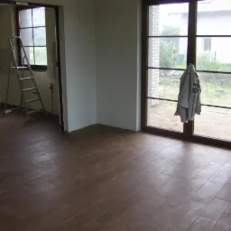 Wnętrze pokoju po ułożeniu brązowych płytek podłogowych imitujących drewno, widoczne drzwi balkonowe z powieszonym płaszczem i drabina w przejściu do innego pomieszczenia.