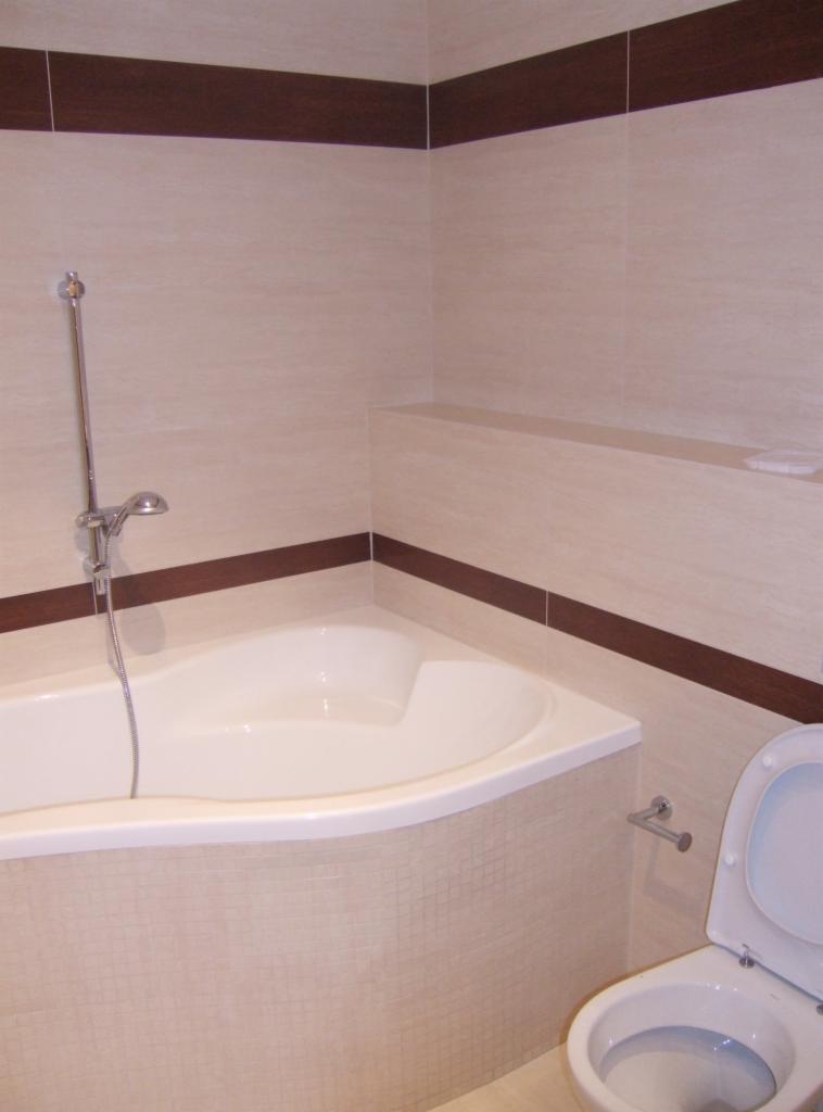 Jasna łazienka z narożną wanną obudowaną mozaiką, toaletą z otwartą klapą i brązowymi akcentami płytek.