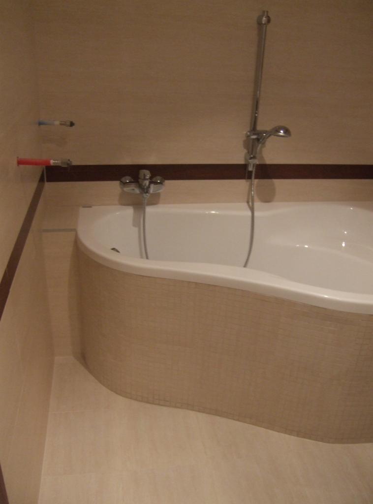 Biała narożna wanna obudowana mozaiką w beżowych odcieniach, chromowana bateria z prysznicem, beżowe płytki na ścianach i podłodze, widoczne rury doprowadzające wodę.