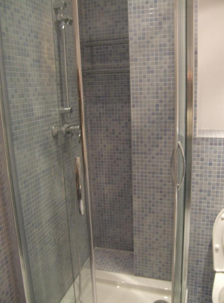 Szklana kabina prysznicowa z chromowanymi okuciami i deszczownicą, wyłożona mozaiką w odcieniach szarości i błękitu, z widoczną częścią toalety po prawej stronie.