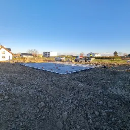 Wylana płyta fundamentowa z widocznymi prętami zbrojeniowymi i elementami szalunku, ekipa budowlana przy samochodzie dostawczym w tle, krajobraz wiejski.