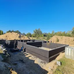 Fundamenty domu jednorodzinnego z deskowaniem, zabezpieczone czarną powłoką, z widocznym zbrojeniem i nasypem ziemi w tle.