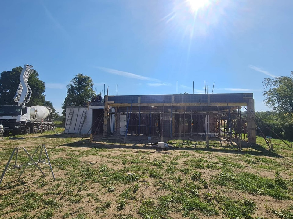 Budowa domu murowanego w trakcie wznoszenia ścian, widoczny betonowóz z pompą do betonu, szalunki i zbrojenia, słoneczny dzień.