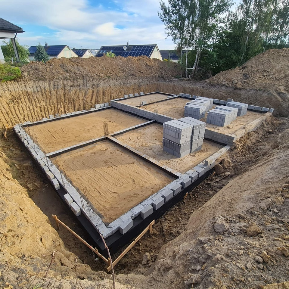 Fundament domu w wykopie, częściowo wymurowany z bloczków betonowych, z widoczną hydroizolacją i piaskiem wewnątrz, przygotowany do dalszych prac budowlanych.