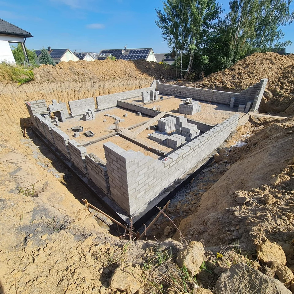 Fundamenty domu z bloczków betonowych, częściowo wymurowane, widoczne z wykopu, z zapasem bloczków na paletach wewnątrz, w tle domy jednorodzinne i drzewa.