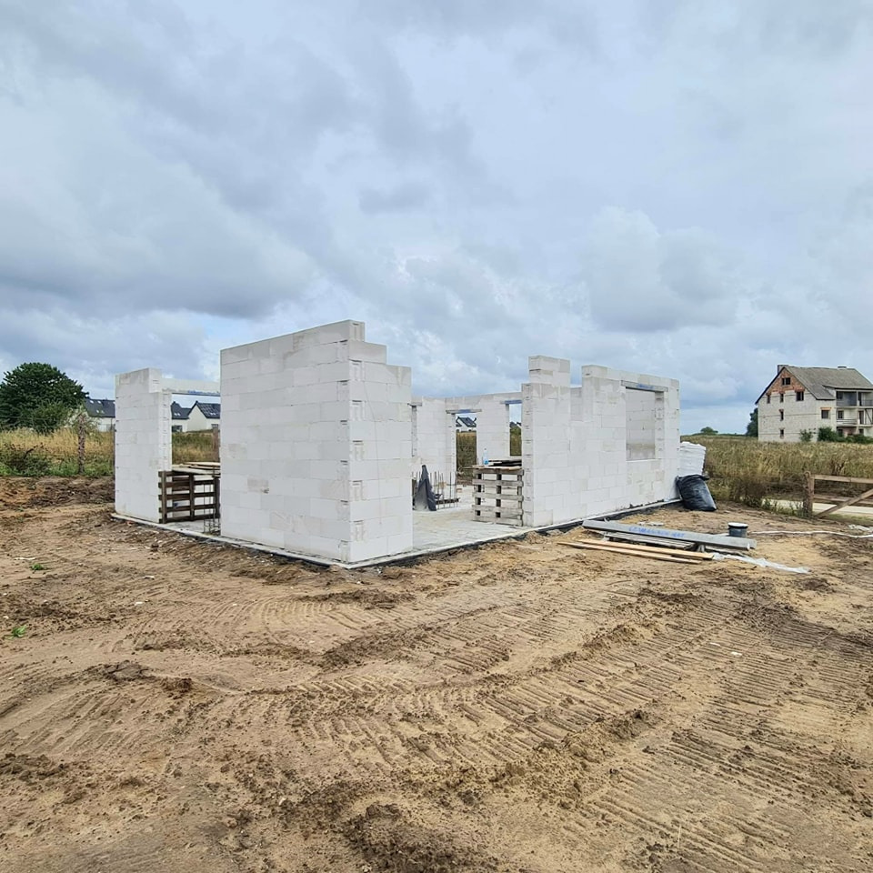 Wznoszenie ścian parterowego domu z bloczków betonu komórkowego, widoczne szalunki okienne i drzwiowe, teren budowy z widocznymi śladami opon, w tle budynek w stanie surowym.