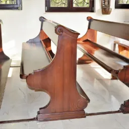 Rząd ciemnobrązowych, drewnianych ławek kościelnych z wysokimi oparciami i półkami na książki, ustawionych na marmurowej podłodze w jasnym wnętrzu.