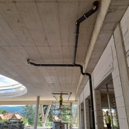 Twój Kąt Sp. z o.o. - Montaż instalacji kanalizacyjnej pod sufitem surowego budynku, widoczni dwaj pracownicy w kamizelkach odblaskowych, jeden na rusztowaniu, drugi stoi obok ściany z betonu komórkowego.