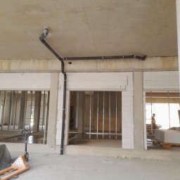 Twój Kąt Sp. z o.o. - Wnętrze surowego budynku w trakcie budowy z widoczną czarną instalacją rurową pod sufitem, wspartą na metalowych obejmach, oraz pustakami na ścianach działowych. Na podłodze widoczne ślady wody...