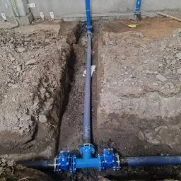 Niebieskie rury instalacji wodnej ułożone w wykopie ziemnym, widoczne złącza skręcane i fragment ściany budynku w tle.