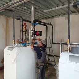 Twój Kąt Sp. z o.o. - Instalator w roboczym kombinezonie montuje rury do białego pieca grzewczego, używając klucza nastawnego; widoczne elementy instalacji, drabina i surowy wystrój pomieszczenia.