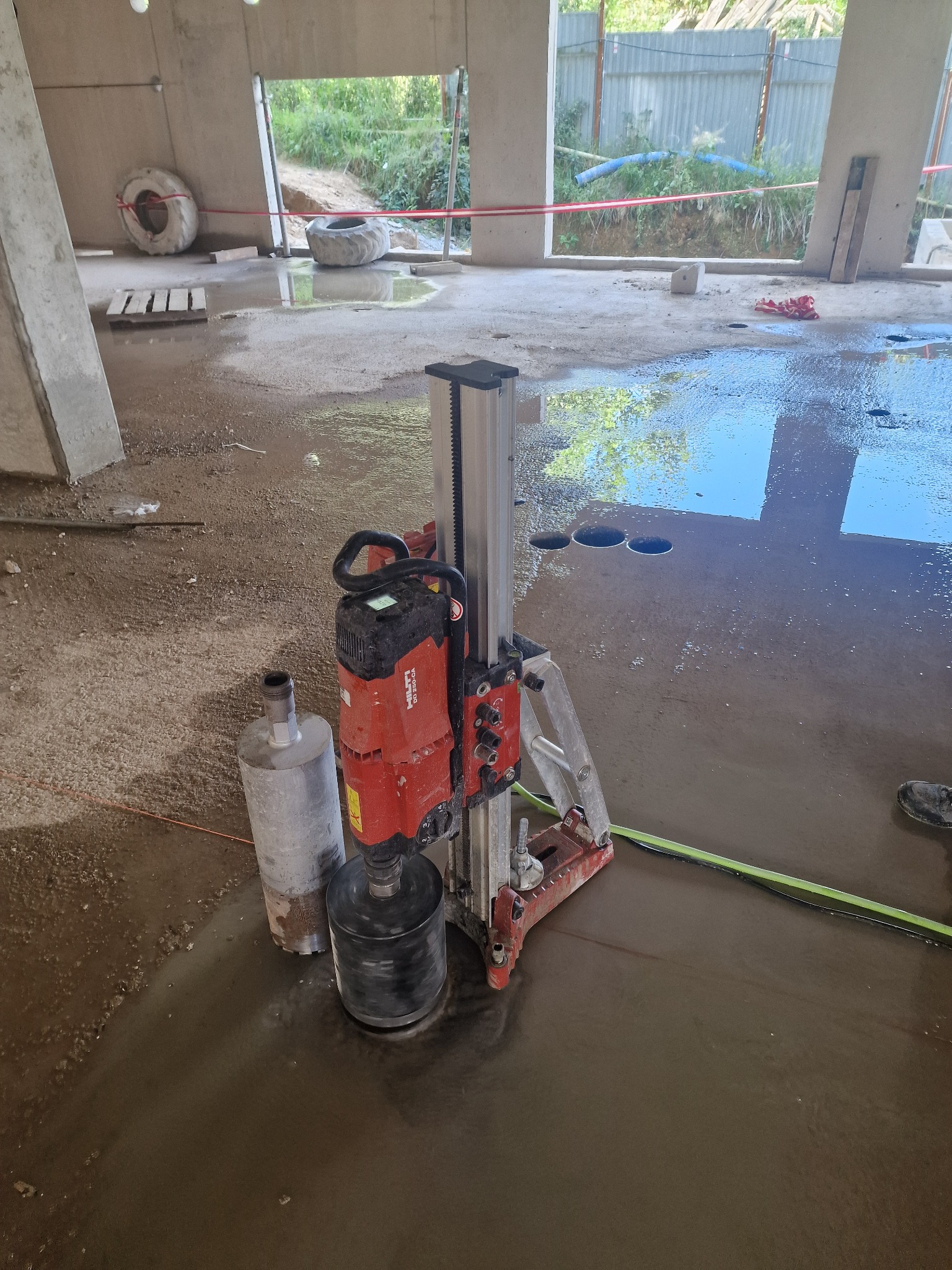 Wiertnica diamentowa Hilti w trakcie pracy na mokro w surowym wnętrzu budynku w Świerzawie, widoczne wywiercone otwory w posadzce i odbicie nieba w wodzie.