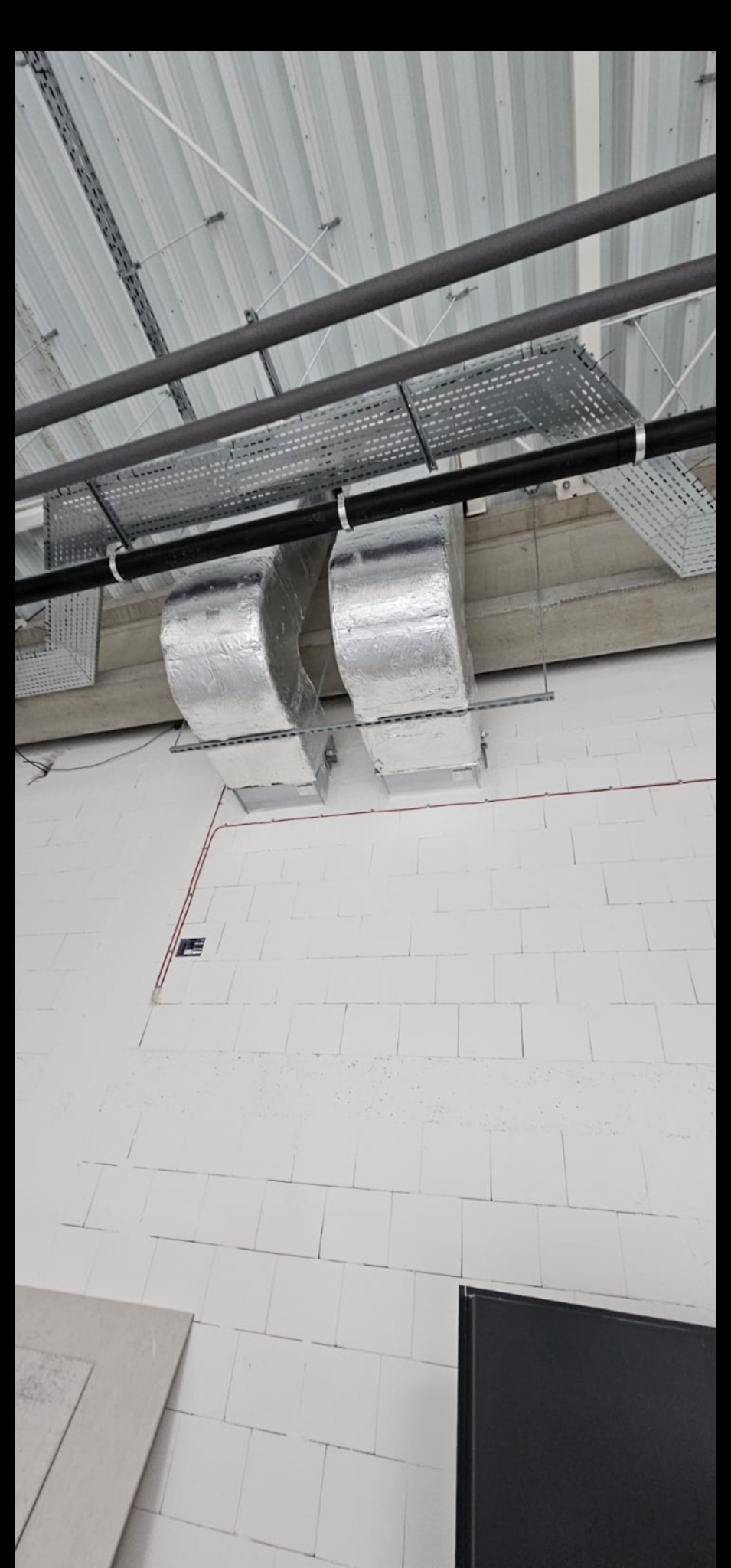 Dwa przewody wentylacyjne w srebrnej izolacji zamontowane pod sufitem z blachy trapezowej, widoczne czarne rury i koryta kablowe, ściana z białych bloczków.