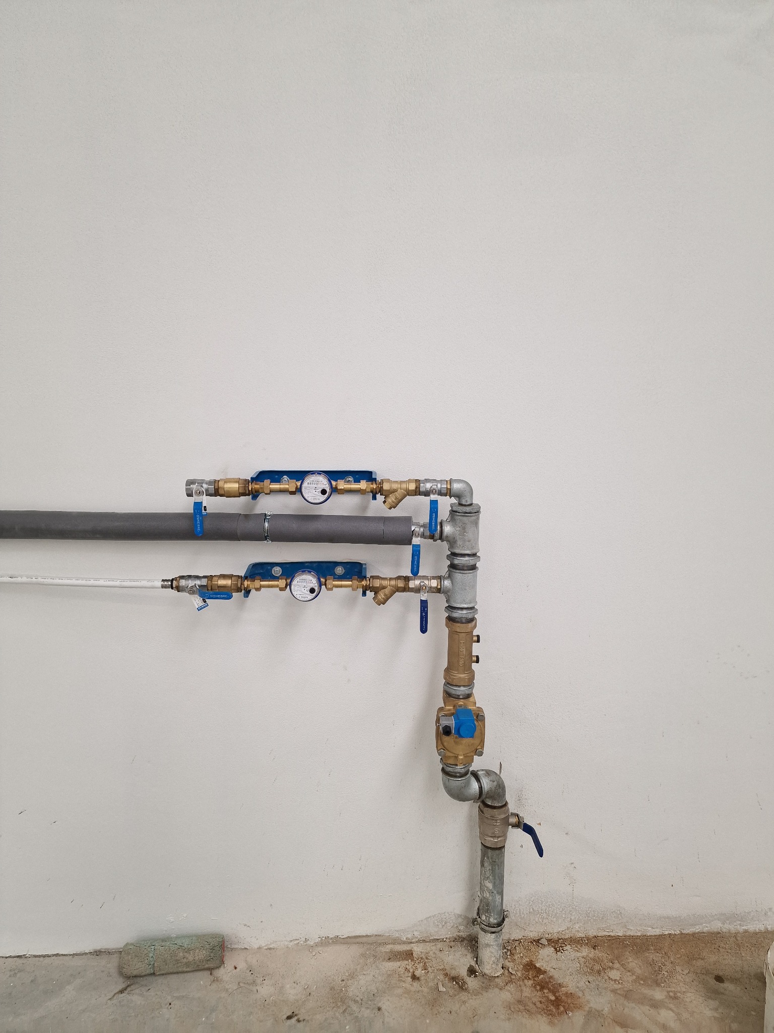 Dwa poziome rzędy rur wodociągowych z licznikami, połączone pionowym odcinkiem rur z zaworami, zamontowane na białej ścianie w surowym stanie.