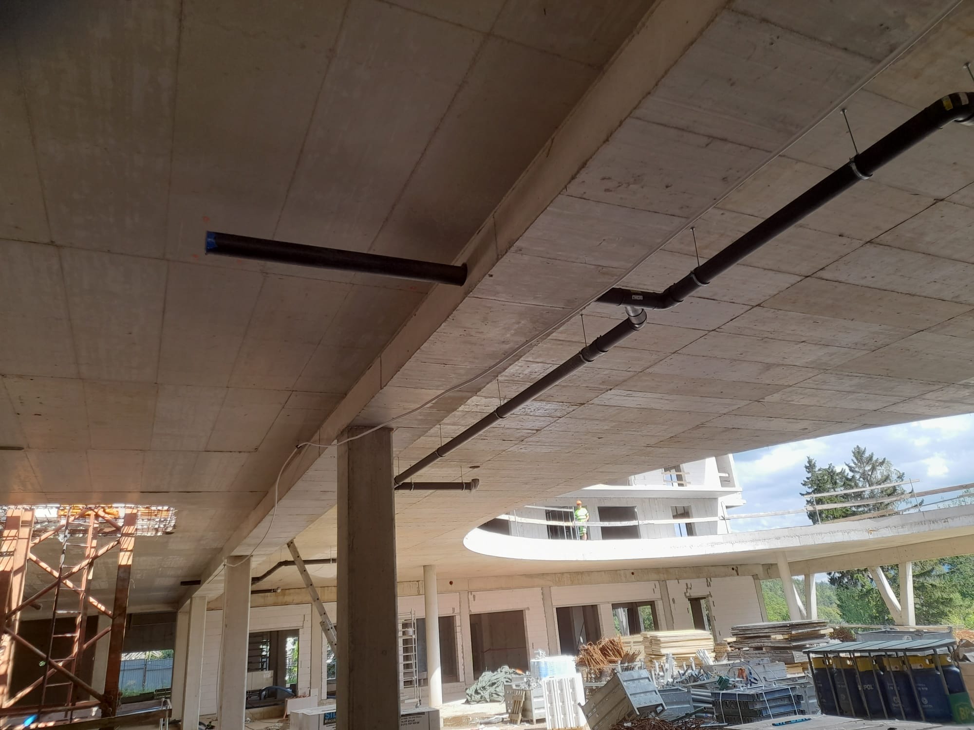 Montaż instalacji kanalizacyjnej pod betonowym stropem budynku w stanie surowym otwartym, widoczne rury PCV mocowane do stropu za pomocą metalowych prętów, w tle budynek w trakcie budowy.