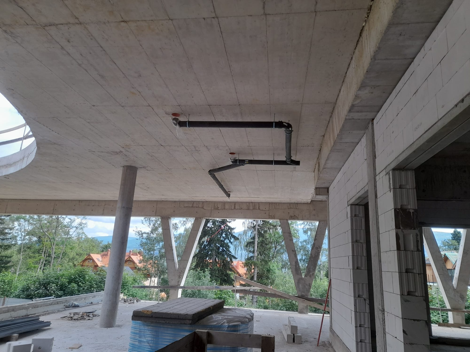 Surowe wnętrze budynku w trakcie budowy z widocznymi czarnymi rurami instalacji podwieszonymi do betonowego stropu, ściany z białych bloczków, widok na zewnątrz na zalesione wzgórza.