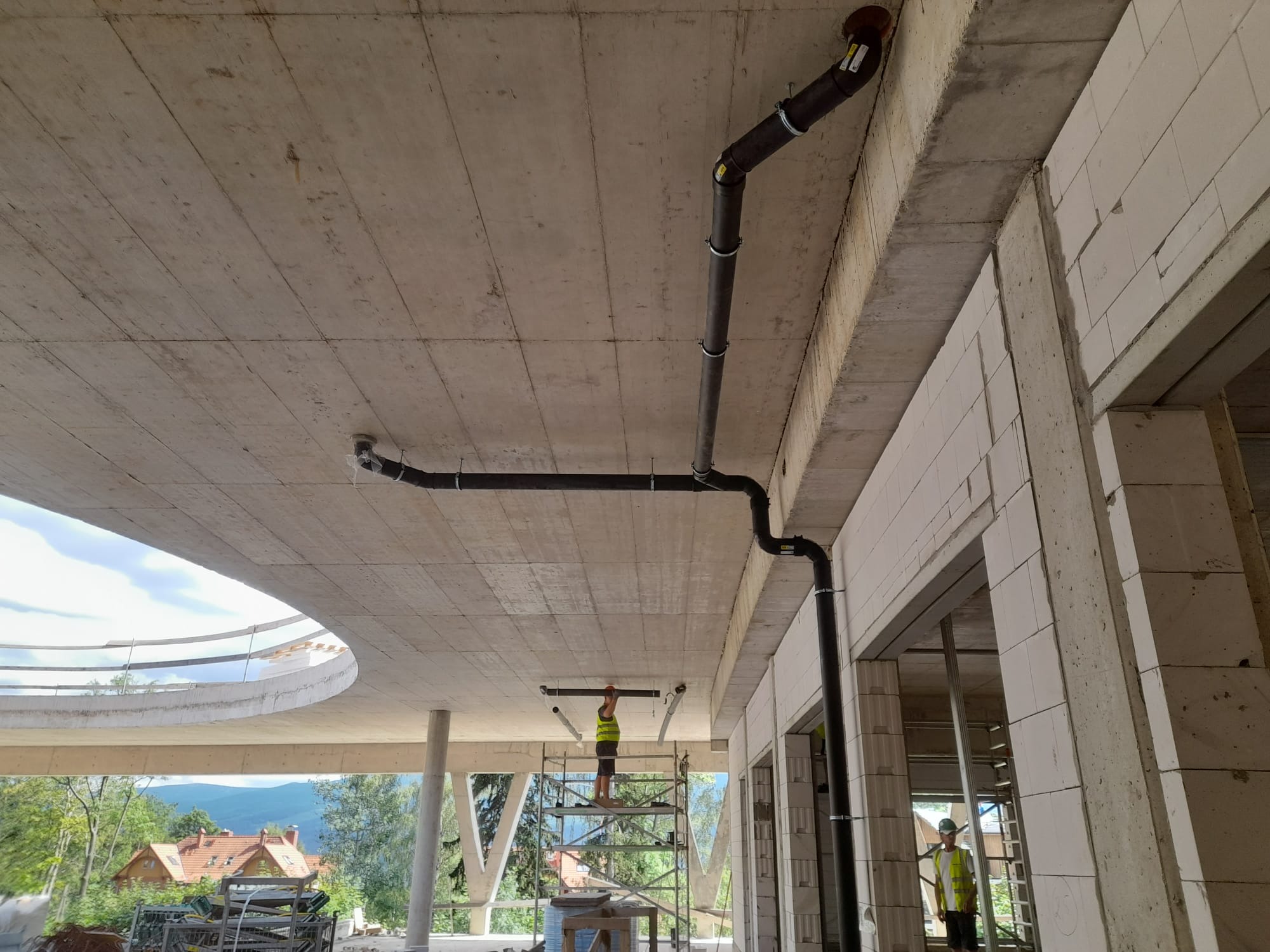 Montaż instalacji kanalizacyjnej pod sufitem surowego budynku, widoczni dwaj pracownicy w kamizelkach odblaskowych, jeden na rusztowaniu, drugi stoi obok ściany z betonu komórkowego.