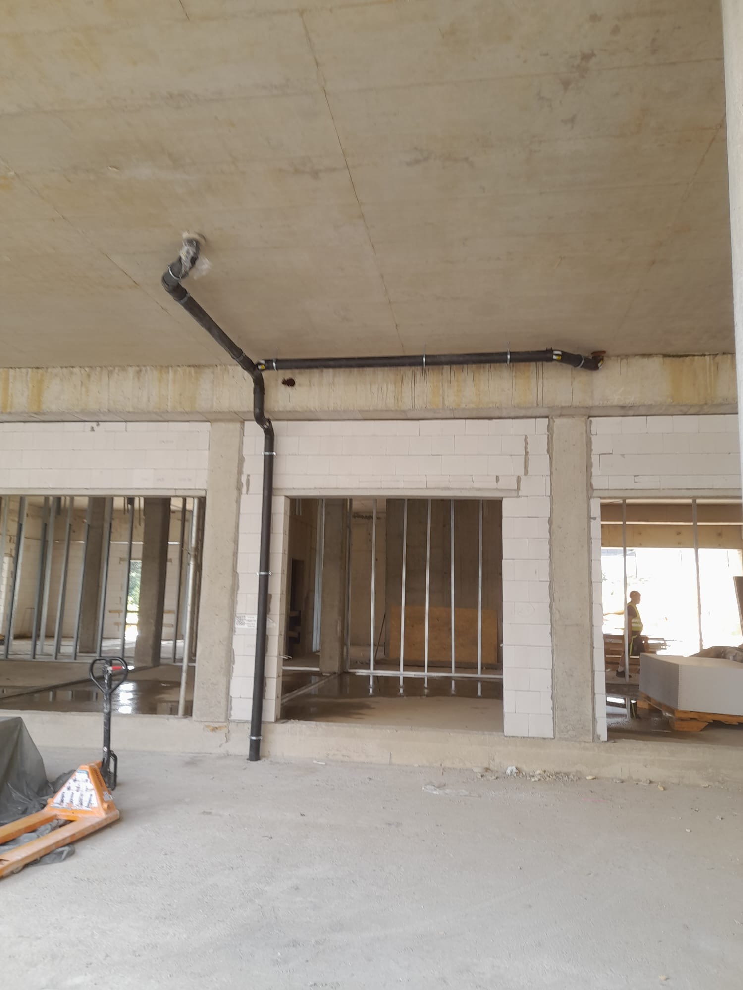 Wnętrze surowego budynku w trakcie budowy z widoczną czarną instalacją rurową pod sufitem, wspartą na metalowych obejmach, oraz pustakami na ścianach działowych. Na podłodze widoczne ślady wody...