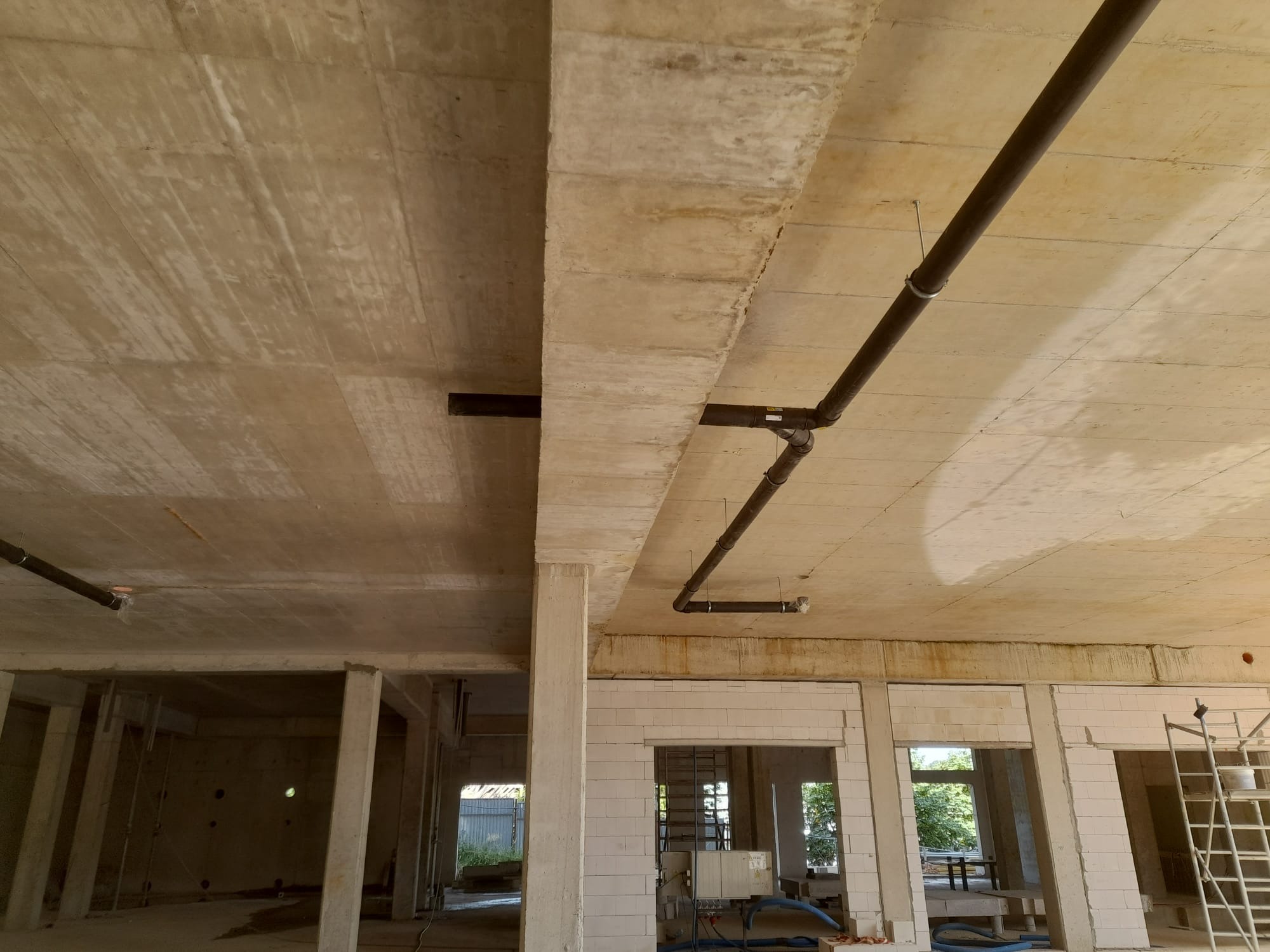 Surowy strop betonowy z widocznymi rurami instalacji podwieszonymi do sufitu, stan surowy budynku z pustakami i oknami bez szyb.