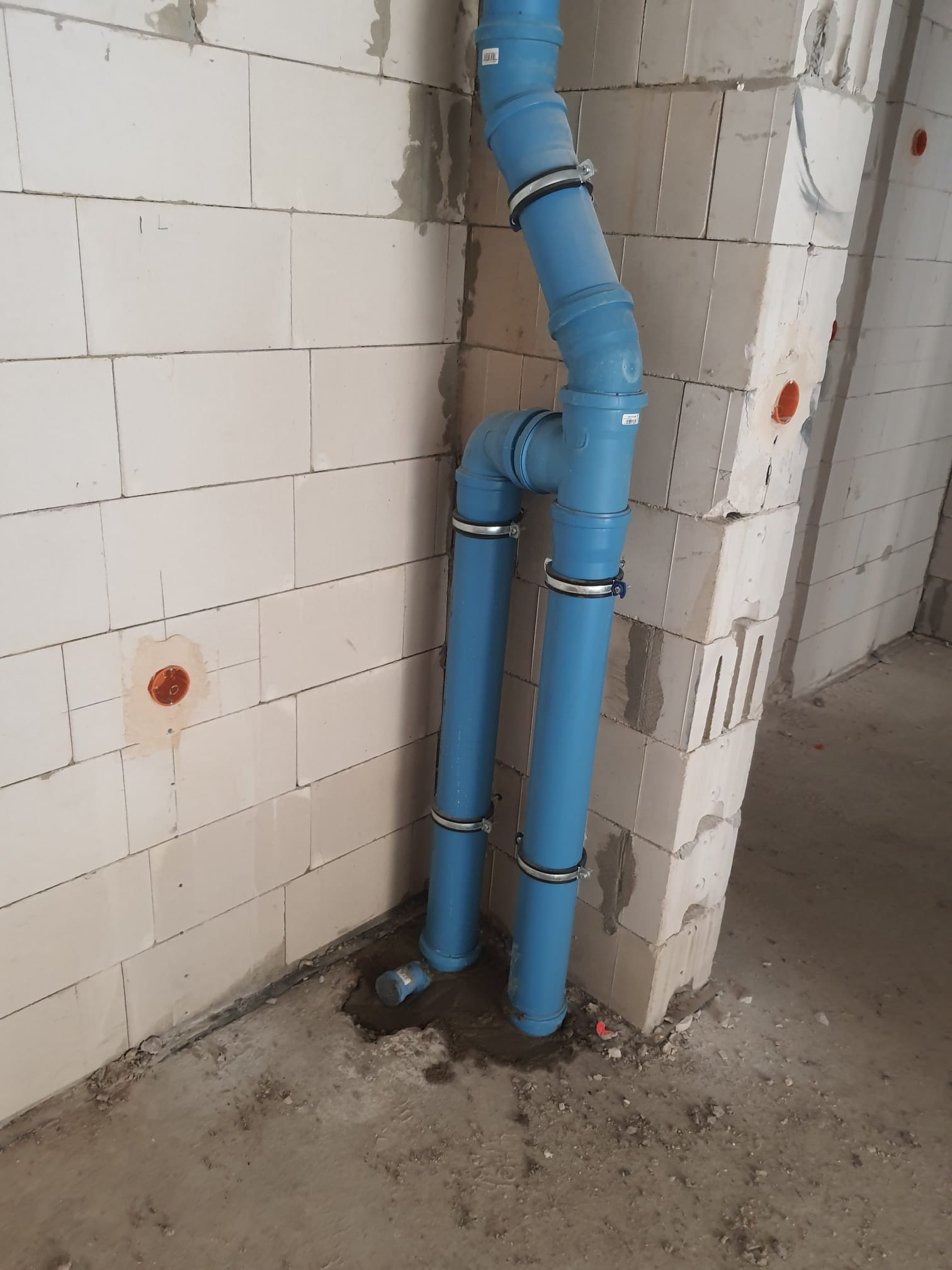 Niebieskie rury kanalizacyjne, mocowane metalowymi obejmami, prowadzone w rogu surowego pomieszczenia z białych bloczków, widoczne otwory na instalacje elektryczne.