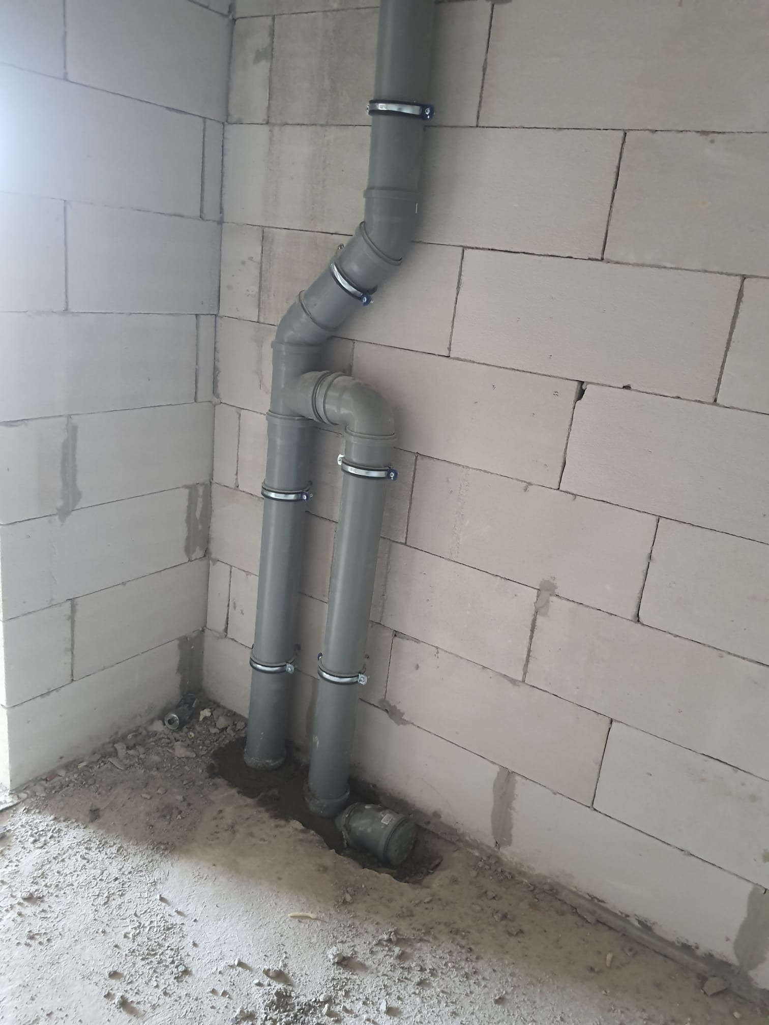 Szare rury kanalizacyjne z metalowymi obejmami zamontowane przy ścianie z betonu komórkowego w surowym stanie.