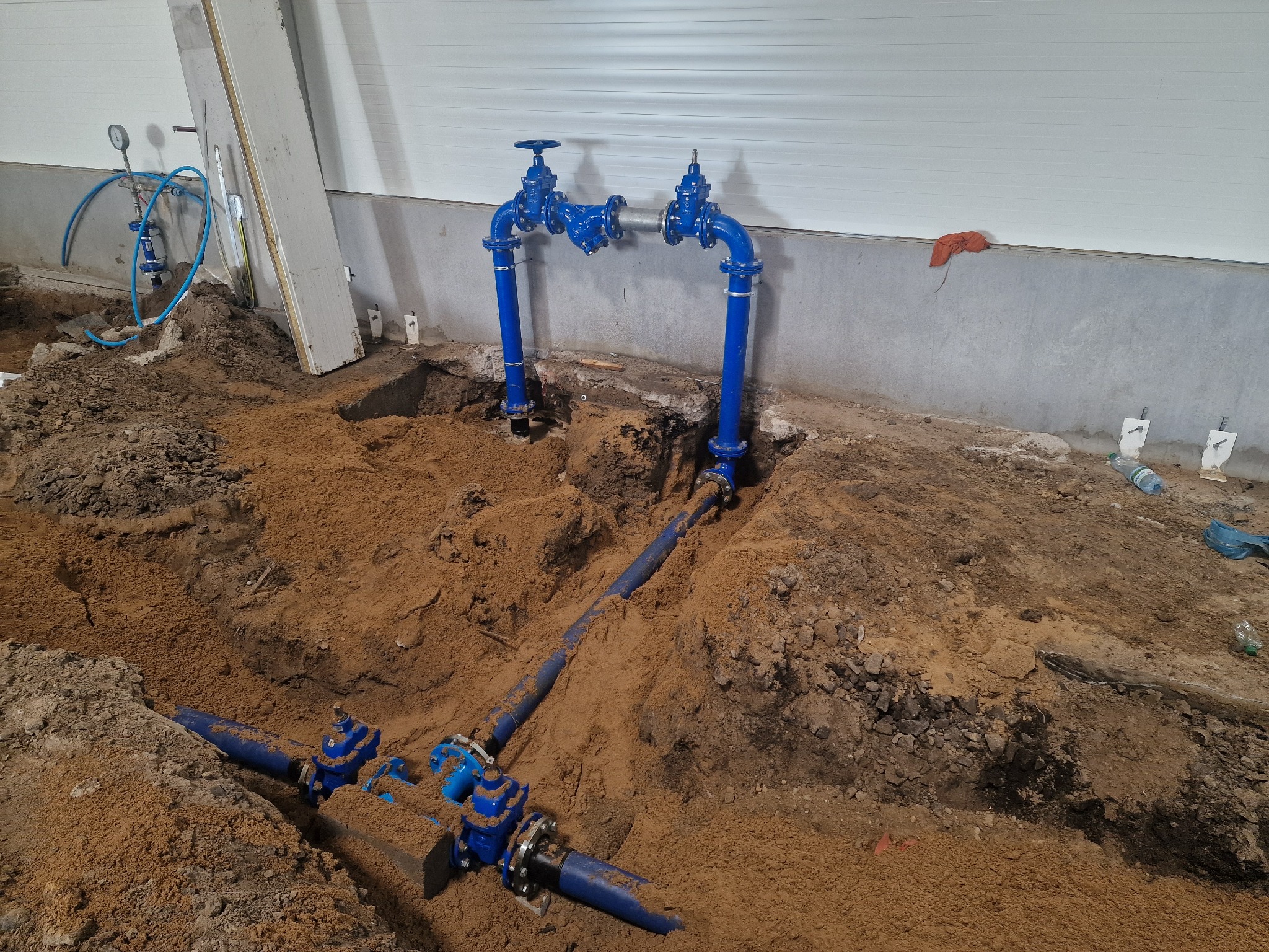 Niebieskie rury z zaworami, częściowo zakopane w ziemi wewnątrz hali, widoczne białe ściany i podłoga w tle.