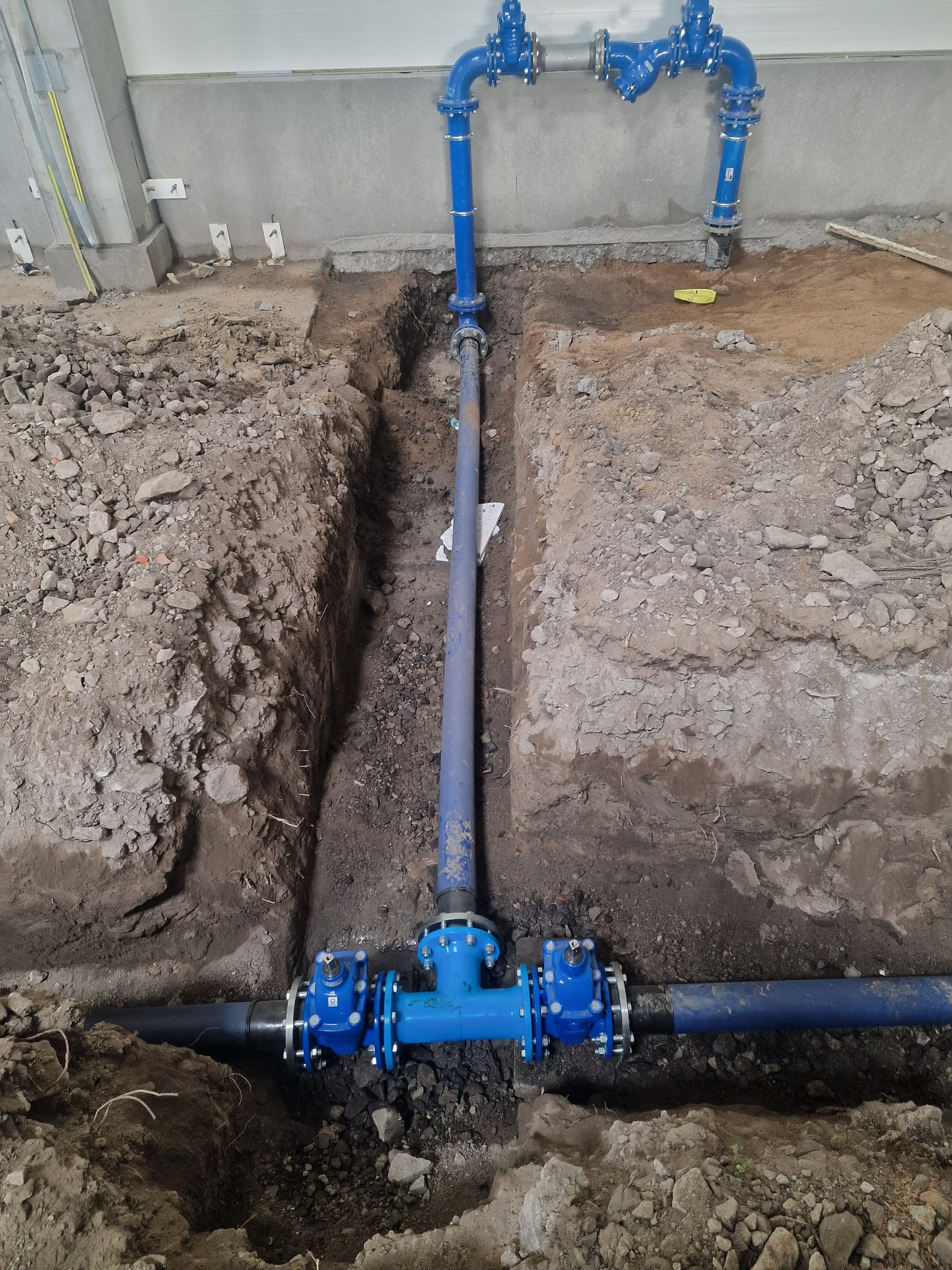 Niebieskie rury instalacji wodnej ułożone w wykopie ziemnym, widoczne złącza skręcane i fragment ściany budynku w tle.