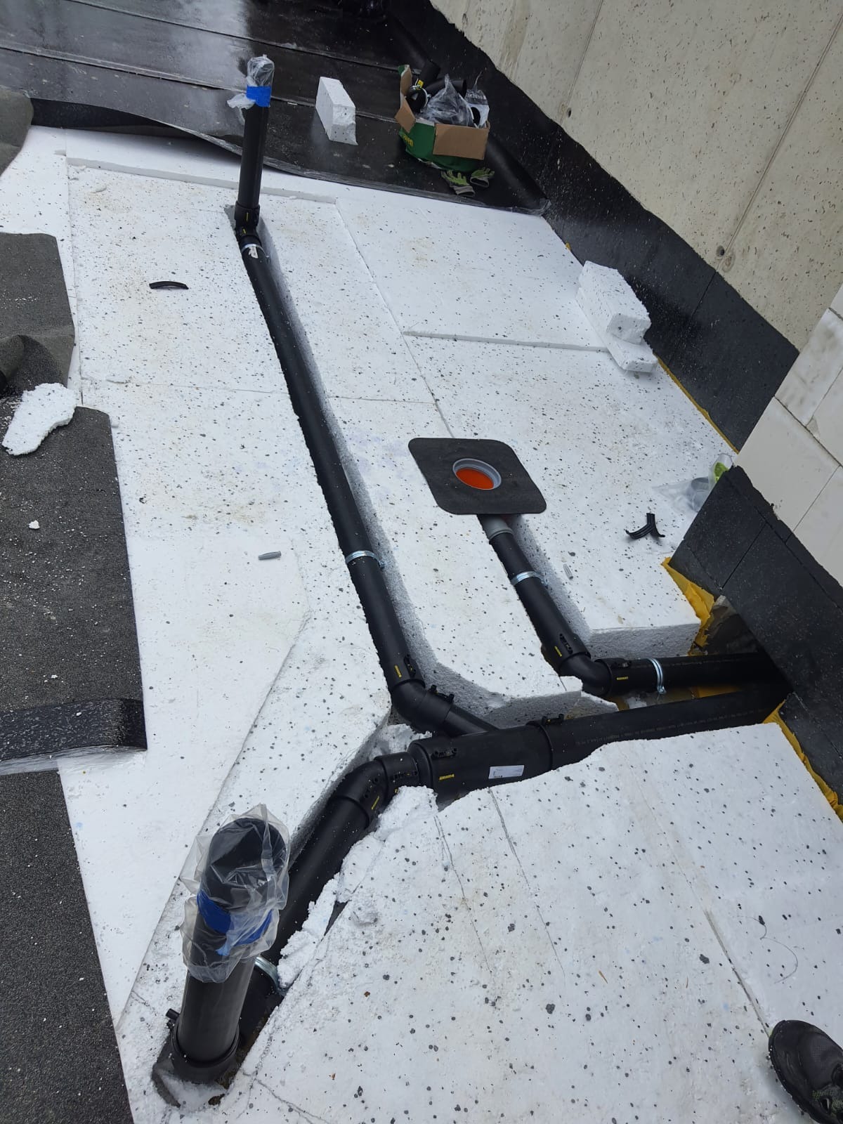 Montaż czarnych rur kanalizacyjnych na białej izolacji styropianowej dachu płaskiego, z widocznym odpływem dachowym i elementami mocującymi.