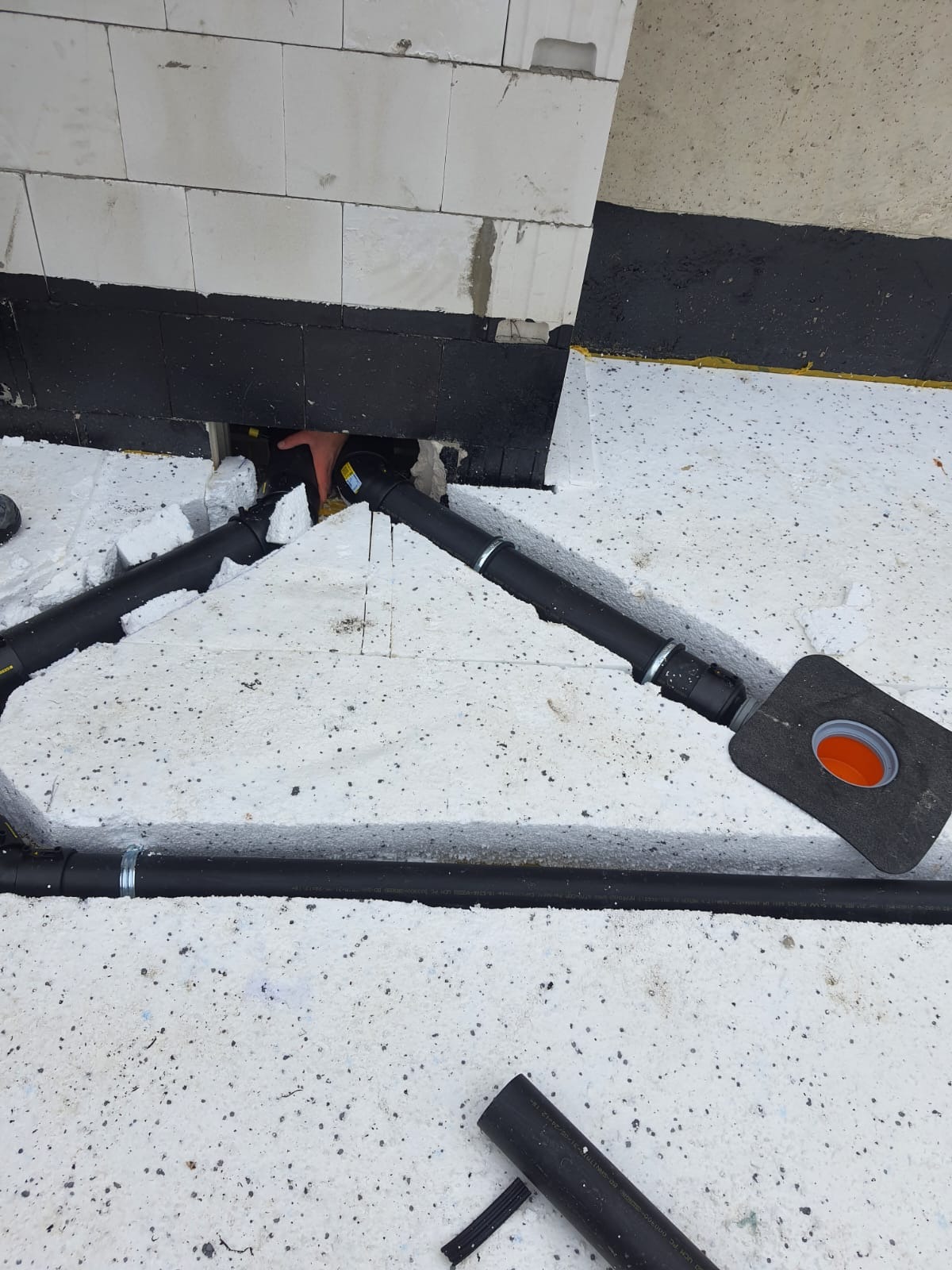 Czarna rura kanalizacyjna z uszczelką pomarańczową w izolacji styropianowej, widoczna dłoń montera w otworze w ścianie z bloczków betonowych.