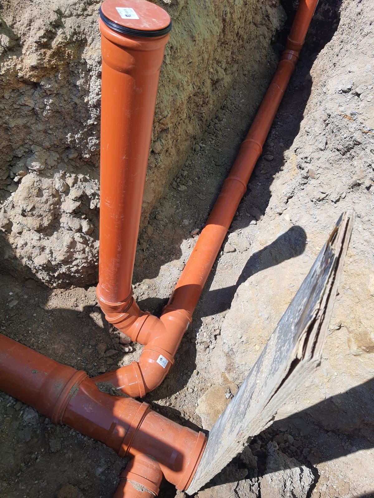 Pomarańczowe rury kanalizacyjne ułożone w wykopie, widoczny fragment łopaty wbijającej się w ziemię.