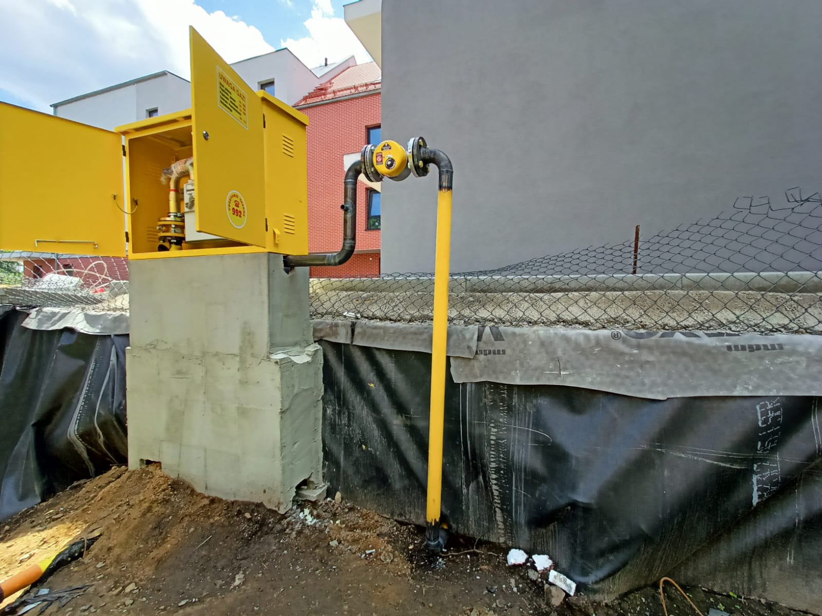 Żółta skrzynka gazowa z otwartymi drzwiczkami, zamontowana na betonowym fundamencie, z rurą gazową w kolorze żółtym, w tle budynki mieszkalne i siatka ogrodzeniowa.