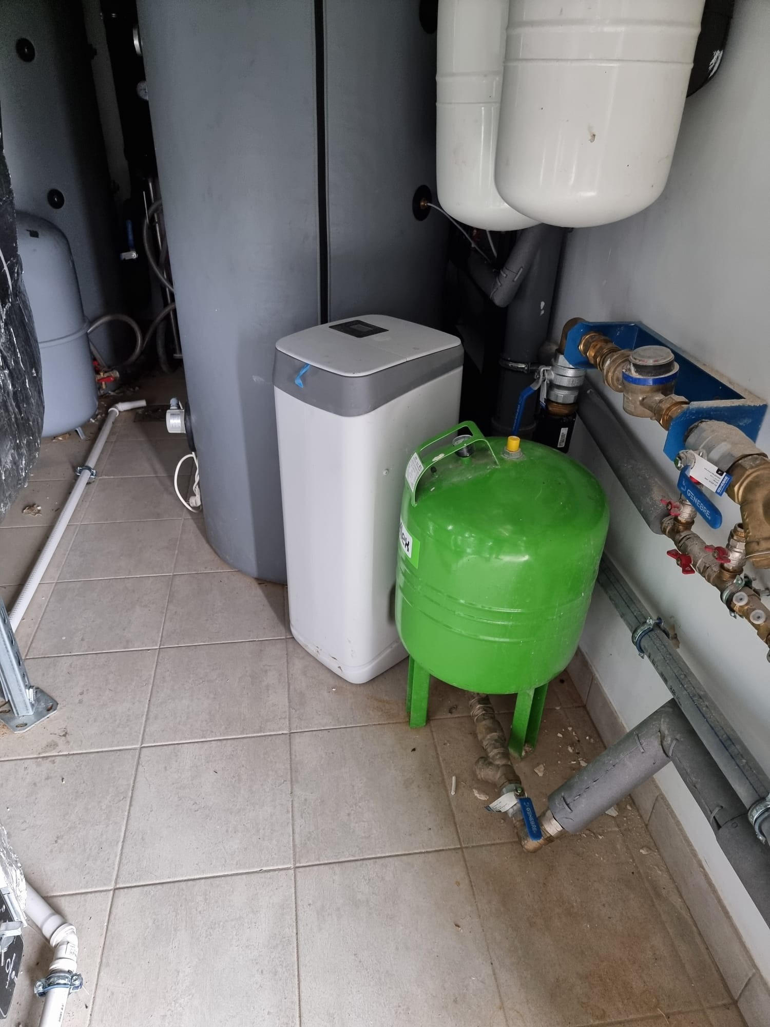 Szary pionowy zbiornik, biały zmiękczacz wody z szarym blatem i zielony zbiornik wyrównawczy na zielonych nóżkach w pomieszczeniu z szarą podłogą i instalacją hydrauliczną.