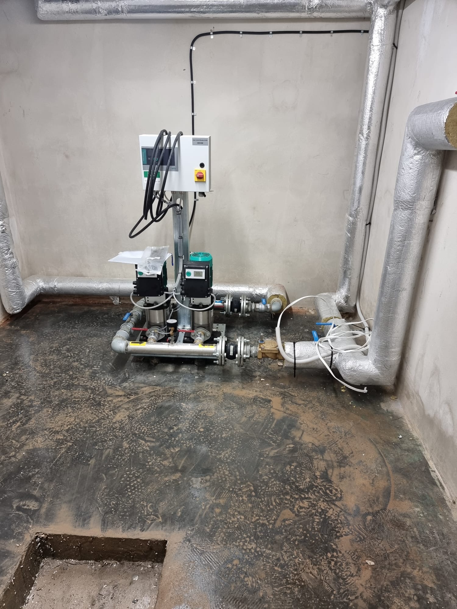 Węzeł hydroforowy z dwiema pompami, sterownikiem i orurowaniem w pomieszczeniu technicznym, widoczne izolowane rury i ślady na podłodze.