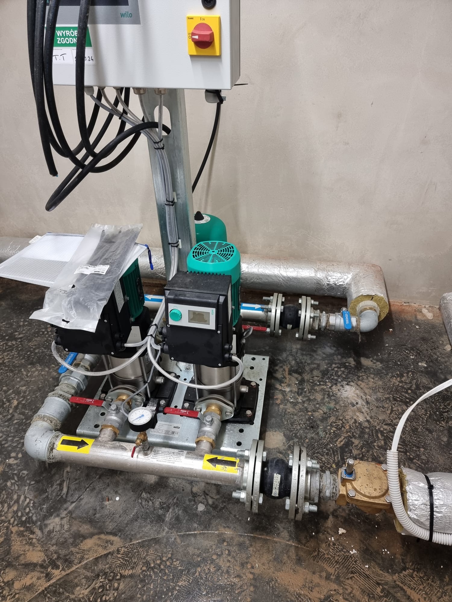 Zmontowany zestaw dwóch pomp obiegowych Wilo na płycie montażowej, połączonych rurami z zaworami i manometrem, widoczne przewody elektryczne, izolacja termiczna rur i panel sterowania na metalowym...