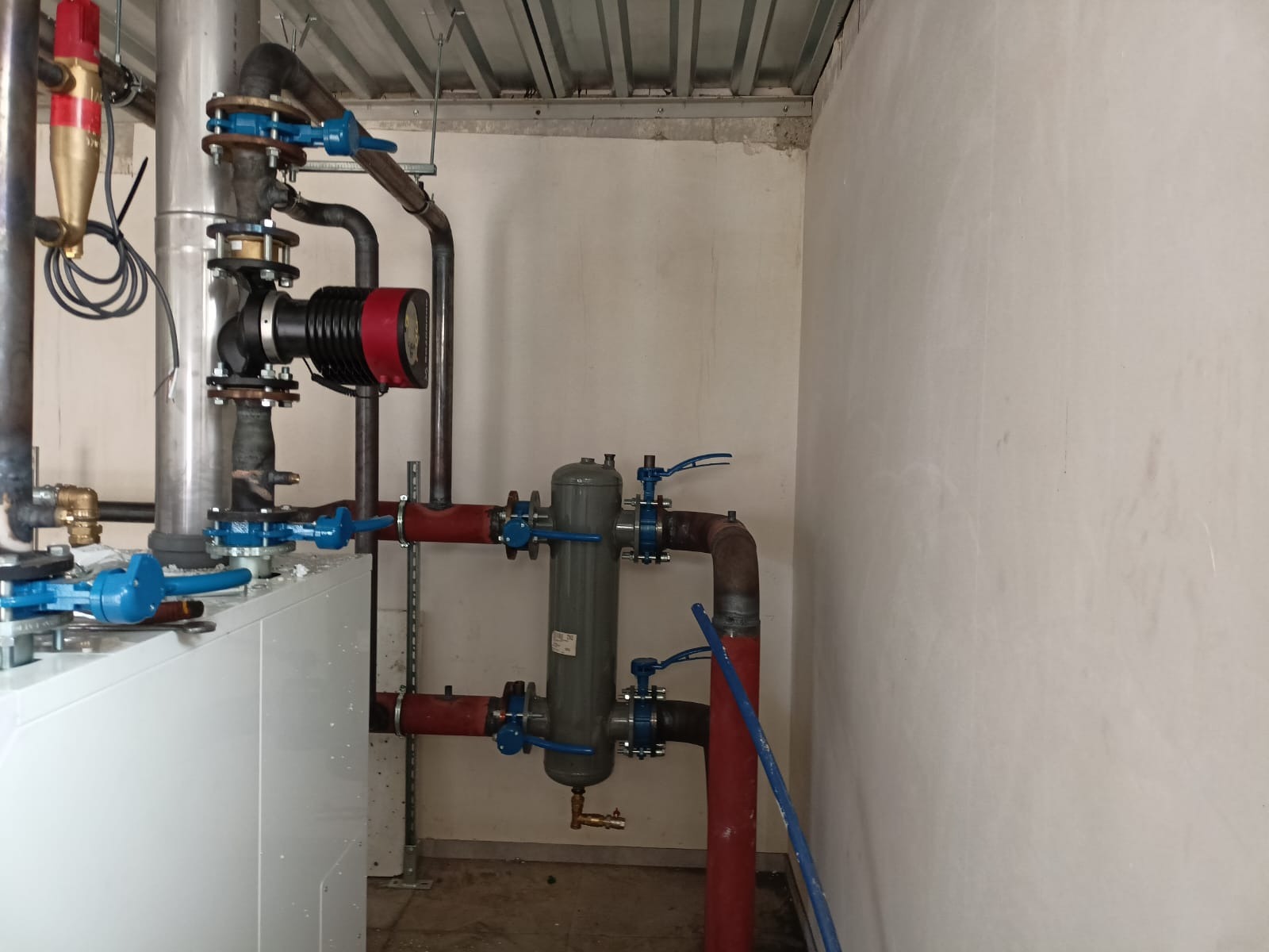 Zmontowana instalacja grzewcza w kotłowni z widocznym piecem, pompą obiegową z czerwonym elementem, zbiornikiem wyrównawczym i licznymi rurami z zaworami o niebieskich uchwytach.