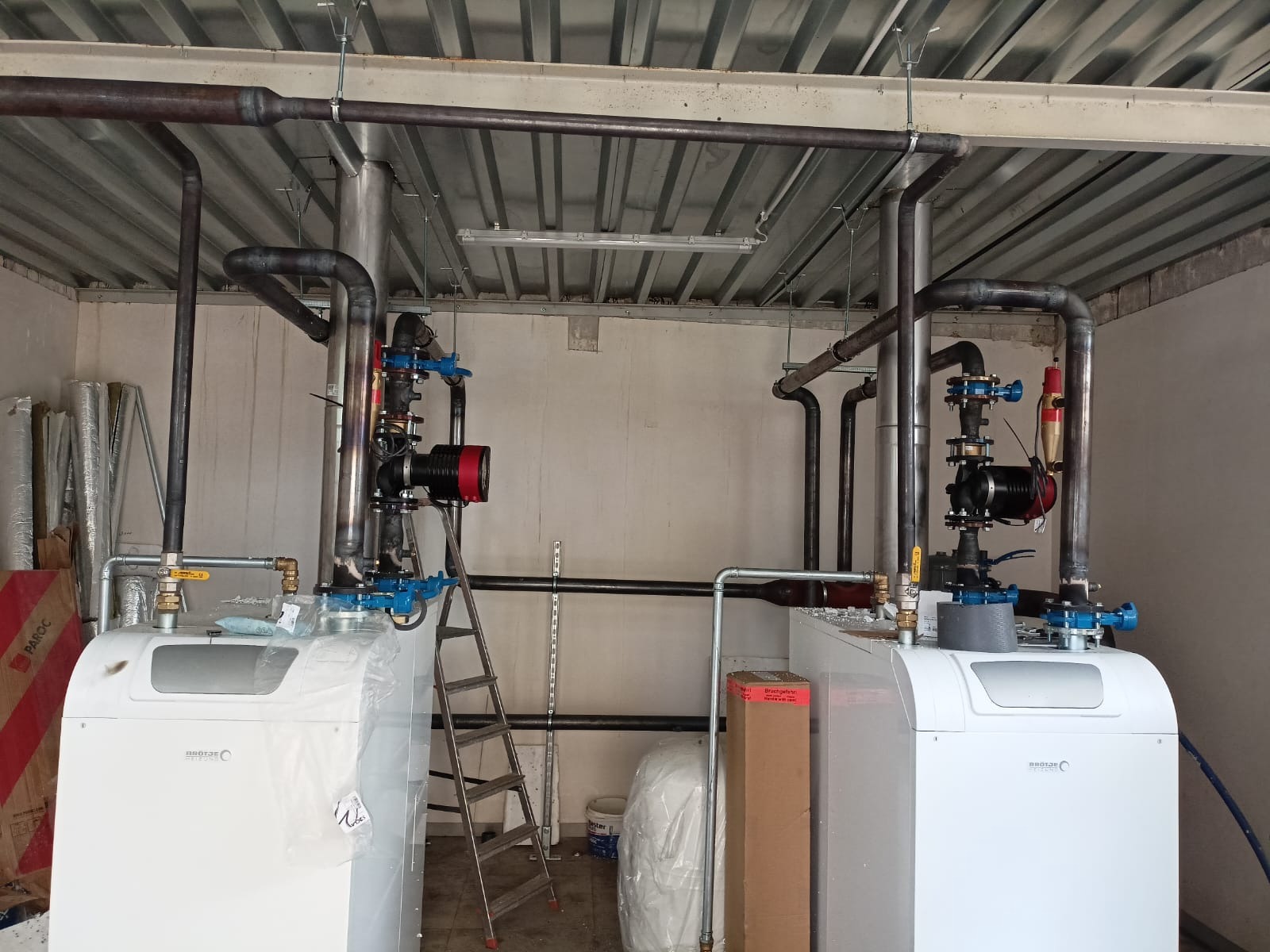 Dwa białe piece grzewcze z podłączonymi rurami, zaworami i pompami obiegowymi w pomieszczeniu technicznym. Widoczna drabina aluminiowa i materiały budowlane.