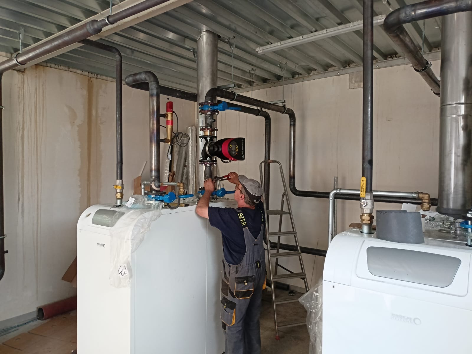 Instalator w roboczym kombinezonie montuje rury do białego pieca grzewczego, używając klucza nastawnego; widoczne elementy instalacji, drabina i surowy wystrój pomieszczenia.