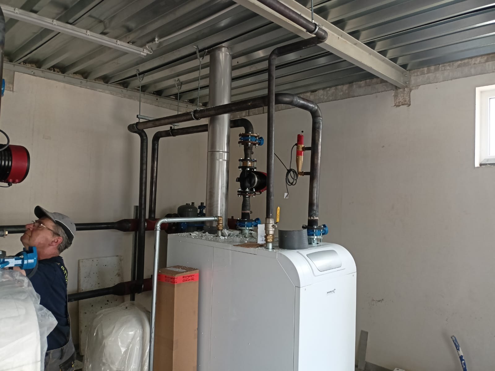 Instalator w okularach, w trakcie montażu kotła grzewczego z licznymi rurami, zaworami i pompą obiegową w kotłowni o surowym wykończeniu.