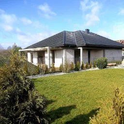 Dom jednorodzinny w stanie surowym zamkniętym z czarną dachówką karpiówką, widok z ogrodu, błękitne niebo z chmurami w tle.