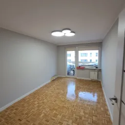 Pusty pokój po remoncie z parkietem w jodełkę, pomalowanymi na szaro ścianami i nowoczesnym oświetleniem sufitowym.