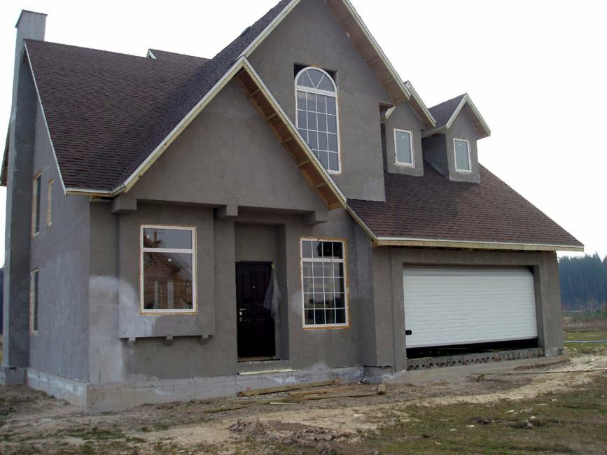 Niedokończona elewacja domu jednorodzinnego z brązowym dachem, widoczne okna z białymi ramami, brama garażowa częściowo otwarta, teren wokół domu niezagospodarowany.