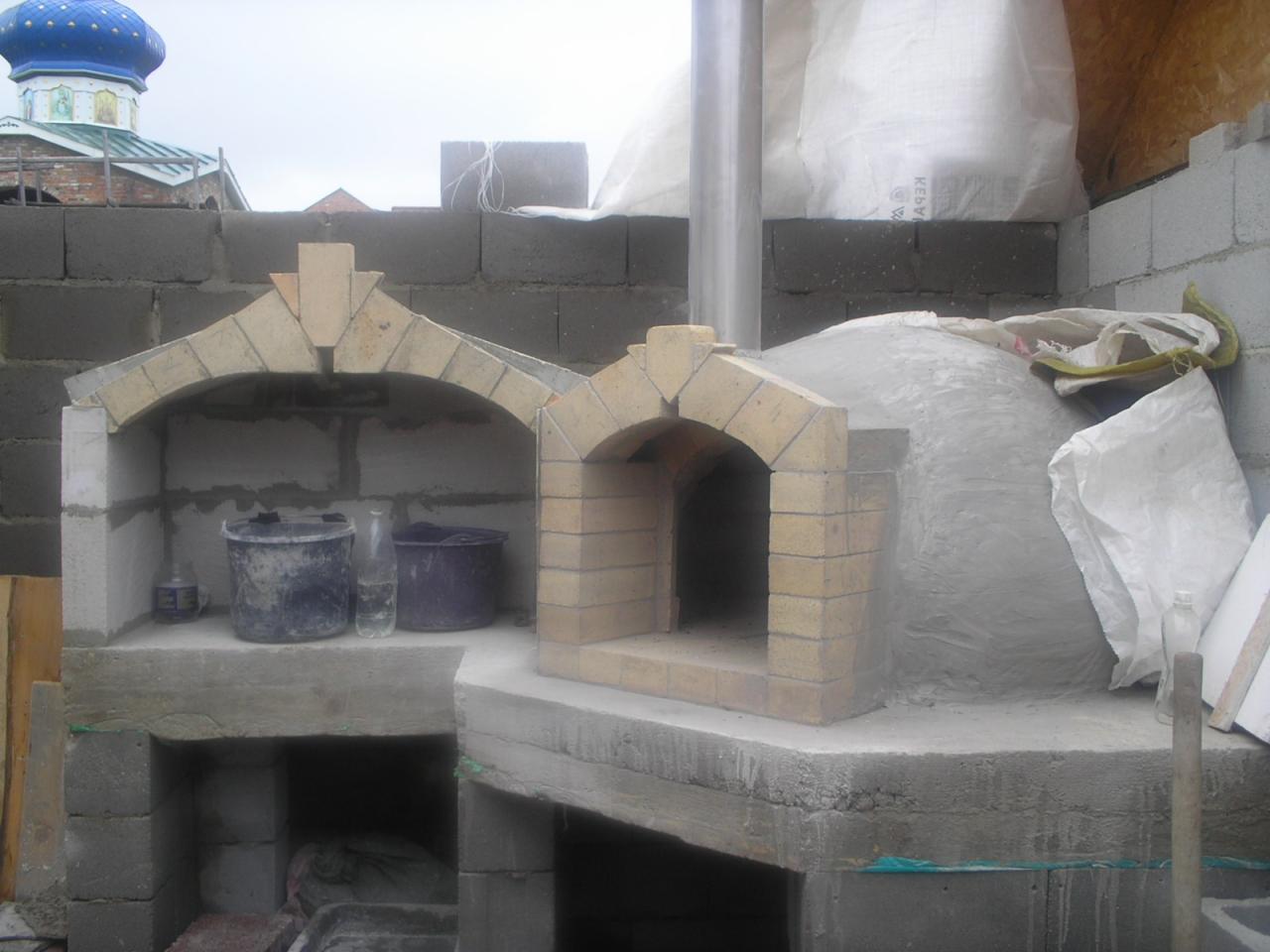Budowa pieca chlebowego z cegły szamotowej z widoczną kopułą w trakcie obkładania zaprawą, obok konstrukcja grilla z cegły, w tle niebieska kopuła cerkwi.
