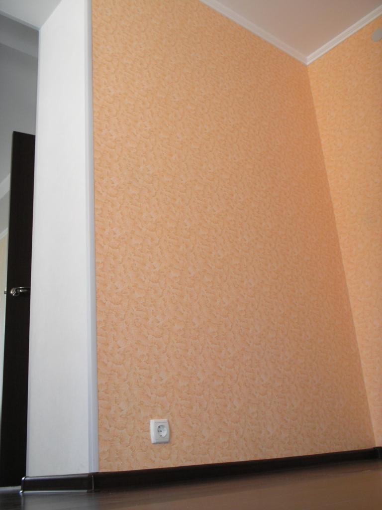 Teksturowana ściana w odcieniach brzoskwini z gniazdkiem elektrycznym, widoczna część futryny drzwi i listwa przypodłogowa.