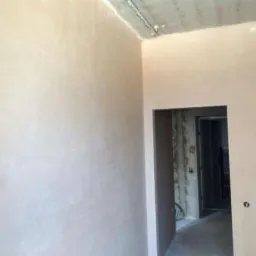 Świeżo nałożony, jasny tynk na ścianach wewnętrznych pomieszczenia, widoczny surowy betonowy sufit i wejście do kolejnego pokoju w trakcie remontu.