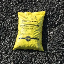 Żółty, foliowy worek 25kg z napisem 'Węglozłom Składy Opału' leży na tle czarnego węgla.