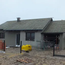 Budowa parterowego domu jednorodzinnego z szarymi ścianami w trakcie montażu więźby dachowej, widoczne rusztowania i materiały budowlane na błotnistym terenie.