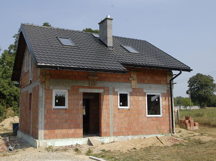 Niedokończony dom murowany z czerwonej cegły z zamontowanymi oknami i dachem z ciemnej blachy trapezowej, widoczne ślady zaprawy i drewniane stemple podtrzymujące nadproża.